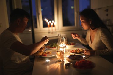 Genç çift evde mum ışığında romantik bir akşam yemeği yiyor, lezzetli yemek ve şarabın tadını çıkarıyor.