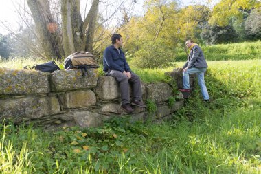 İki erkek yürüyüşçü, güneşli bir doğa ortamında, taş bir duvarda oturup konuşuyorlar.
