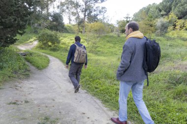 İki erkek turist, güzel bir doğal parkın verimli yeşillikleriyle çevrili toprak bir yolda sakin bir şekilde yürüyorlar.