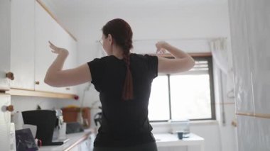 Kızıl saçlı kadın gündelik giysiler giyip mutfakta tek başına dans ediyor.