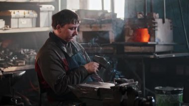 Demirci atölyesinde sigara içerken metal bir parçayı inceliyor.