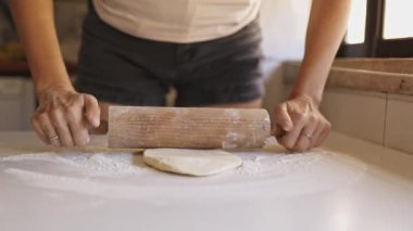 Kadın ahşap bir oklava kullanarak hamuru ustalıkla un kaplı bir zeminde düzleştirir ve pişirmeye hazırlar.