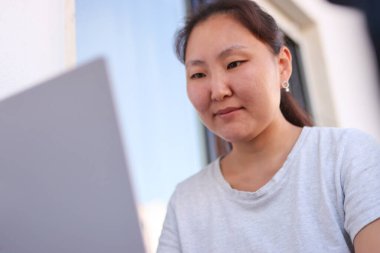 Genç Asyalı kadın dizüstü bilgisayarla uzaktan çalışıyor, ofisinin balkonundaki temiz havanın tadını çıkarıyor.