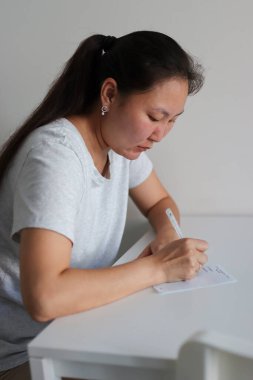 Asyalı bir kadın not defterine kalemle not alıyor, parlak bir odada beyaz bir masada oturuyor.