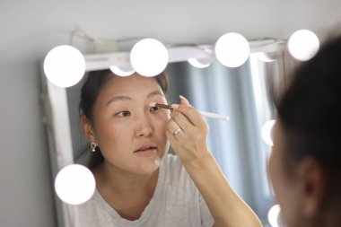 Işıklı aynanın önünde fırça kullanarak göz farı süren Asyalı kadın.
