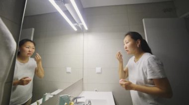 Asyalı bir kadın modern bir banyoda aynaya bakarak yüz kremi sürüyor.