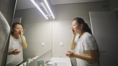 Yüzüne dikkat eden Asyalı kadın banyo aynasının önünde krem sürüyor.