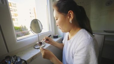 Asyalı bir kadın fırçayla yüzüne fondöten sürüyor.