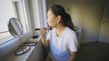 Asyalı bir kadın yüzüne fırça ve yatak odasının penceresinin yanındaki küçük bir ayna kullanarak makyaj yapıyor.