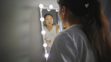 Işıklı aynanın önünde yüzüne makyaj yapan konsantre genç bir kadın.