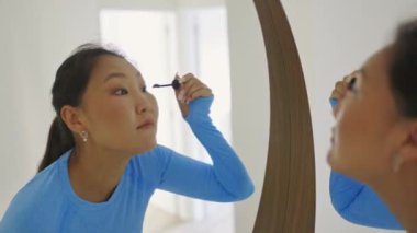 Güzel, genç Asyalı bir kadın maskara sürüyor. Yuvarlak aynada kendine bakıyor.