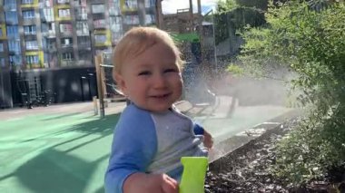 Neşeli çocuk bahçe bitkilerini ilaçlamak için renkli bir su tabancası kullanıyor. Şehir ortamında güneşli bir günün tadını çıkarıyor.