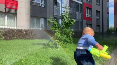 Meraklı çocuk arka bahçedeki bir fıskiyenin yarattığı gökkuşağının yanında su tabancasıyla oynuyor.