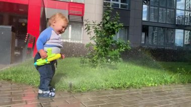 Küçük çocuk su tabancasıyla oynayıp bahçe fıskiyesiyle etkileşime geçiyor.
