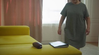 Yaşlı kadın kanepeye yaklaşıyor, oturuyor, gözlük kutusunu açıyor, gözlüklerini çıkarıp takıyor.