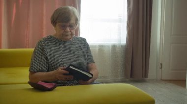 Sarı bir kanepede oturan yaşlı kadın kapıyı açar ve bir kitap okur.