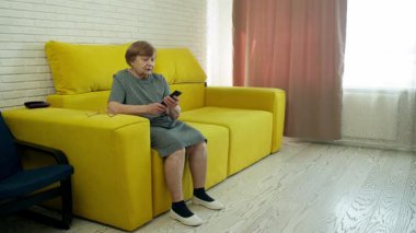 Sarı bir kanepede, elinde akıllı bir telefonla dinlenen yaşlı bir kadın.