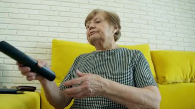 Üst düzey kadın uzaktan kumanda kullanıyor, sarı kanepeden televizyon kanallarında geziniyor.