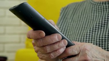 Uzaktan kumandayı elinde tutan, televizyon izleyen, sarı koltukta oturan yaşlı bir kadın.