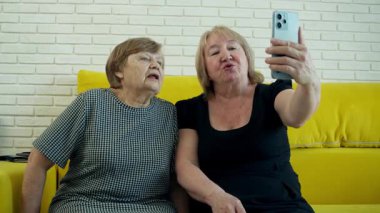 İki yaşlı kadın sarı bir kanepede oturup akıllı telefonlarıyla selfie çekiyorlar.