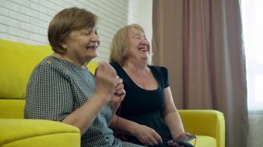 İki son sınıf öğrencisi kadın kulaklıklarını paylaşıyor, akıllı telefondan müzik dinliyor ve sarı bir kanepede birlikte gülüyorlar.