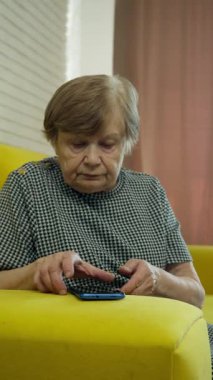 Yaşlı kadın sarı bir koltukta oturuyor, akıllı telefonunu kullanıyor.