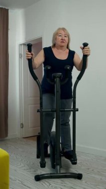 Oturma odasında eliptik makine üzerinde egzersiz yapan, aktif yaşam tarzını sürdüren kararlı yaşlı kadın.