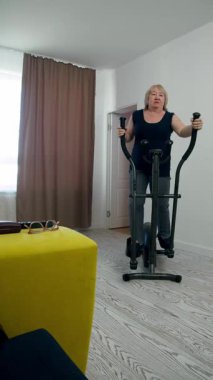 Oturma odasında eliptik çapraz antrenör üzerinde egzersiz yapan yaşlı bir kadın.