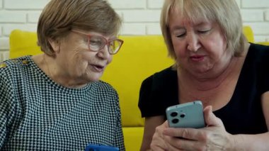 İki yaşlı kadın sarı bir kanepede oturuyor, akıllı telefona bakıyor ve içeriğini tartışıyorlar.