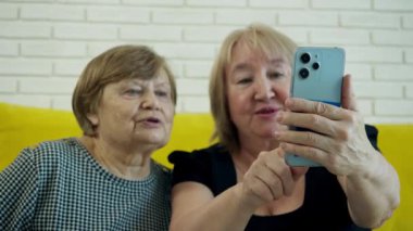 İki yaşlı kadın akıllı telefonlarıyla selfie çekiyor, gülümsüyor ve eğleniyorlar.