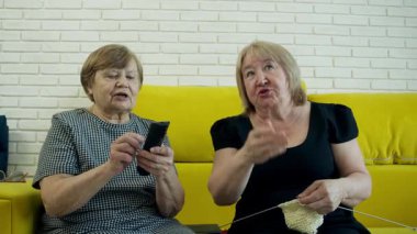 İki son sınıf öğrencisi kadın televizyon izlemeyi ve birlikte örgü örmeyi seviyor, rahat sarı bir kanepede dinlenme anlarını paylaşıyorlar.