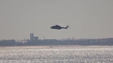 Askeri helikopter arka planda sanayi binaları olan bir kıyı bölgesinin üzerinde uçuyor.