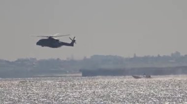 Denizin üzerinde uçan askeri helikopter, bir uçak gemisi ve bir motorlu tekne yakınında.