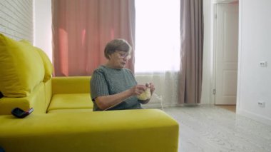 Kıdemli kadın hobisinin tadını çıkarıyor, sarı yün örerek oturma odasındaki rahat sarı kanepeye oturuyor.