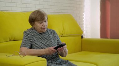 Televizyon izleyen yaşlı bir kadın, sarı kanepede kanal değiştiriyor.