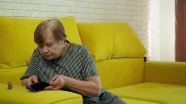 Sarı bir kanepede oturan yaşlı kadın akıllı telefonunu kullanarak bir telefon görüşmesi yapıyor.