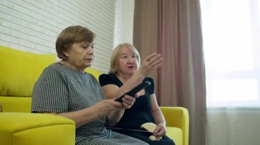 İki yaşlı kadın sarı bir kanepede oturup televizyon seyrediyor ve örgü örüyor.