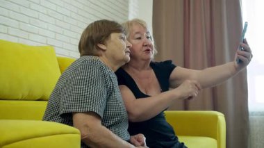 İki yaşlı kadın sarı bir kanepede oturup akıllı telefonlarıyla selfie çekiyorlar.
