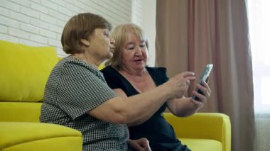 İki yaşlı kadın sarı bir kanepede oturuyorlar, video görüşmesi için akıllı telefon kullanıyorlar, ekranı gösteriyorlar ve el sallıyorlar.