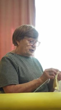 Odaklanmış yaşlı bir kadın beyaz yün bir elbise örüyor, bir pencerenin yanında rahatça oturuyor.