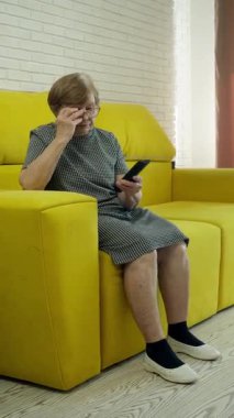 Üst düzey kadın akıllı telefon ve televizyon arasında geçiş yapıyor, sarı bir kanepede çeşitli duygular yaşıyor.