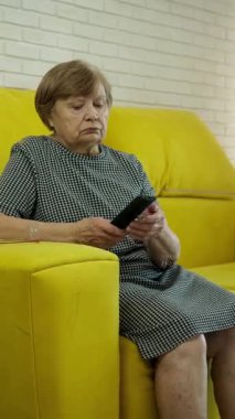 Sarı koltukta oturan yaşlı kadın, elinde uzaktan kumandayla.