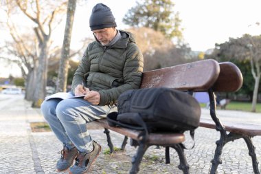 Olgun bir adam parktaki bankta otururken defterine not alıyor.