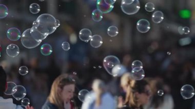 Canlı bir şehir ortamının arka planında canlı sabun kabarcıkları yüzer ve patlar. Tuhaf bir atmosfer yaratır.