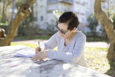 Park masasındaki deftere yazı yazan gözlüklü son sınıf öğrencisi bir kadın, keyifli havanın tadını çıkarıyor.