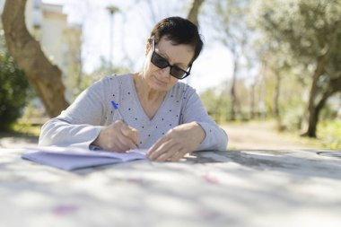 Park masasındaki deftere yazan güneş gözlüğü takan yaşlı bir kadın.