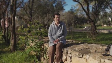 Günlük giysiler giyen kadın parkta alçak taş duvarda oturur, konuşur ve el kol hareketi yapar.