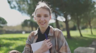 Parkta elinde defterle gülümseyen genç bir kızın portresi.