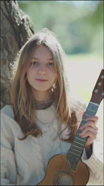 Genç bir kadın ukulele tutuyor, saçlarını düzeltiyor, ve güneşli bir günün tadını çıkarıyor, bir ağacın altında, huzurlu ve müzikal bir anı yakalıyor.