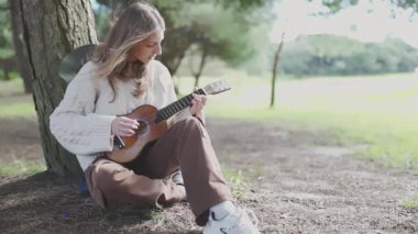 Parktaki bir ağacın altında ukulele çalan genç bir kadın.